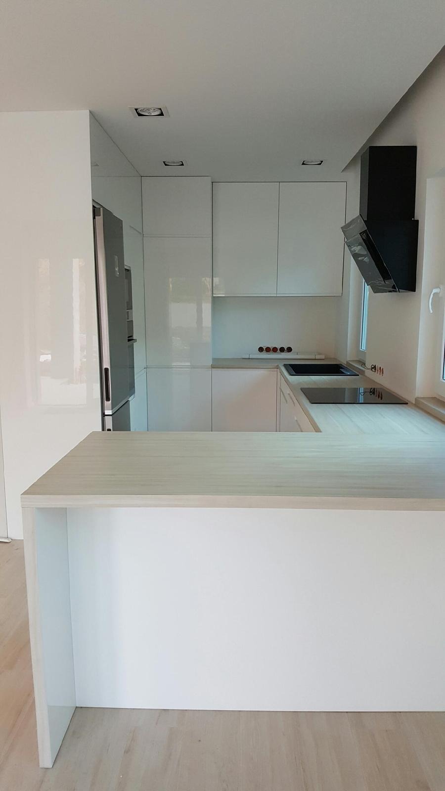 Nowoczesna, minimalistyczna kuchnia z białymi, lakierowanymi frontami, wyspą z jasnym blatem drewnopodobnym, czarnym okapem i wbudowaną płytą grzewczą, oświetleniem punktowym i widocznymi gniazdkami.