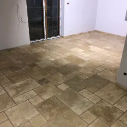 Wykończona podłoga z płytek imitujących kamień w nowym pomieszczeniu, widoczne ślady po instalacji elektrycznej na ścianach, okno z naklejoną folią ochronną.