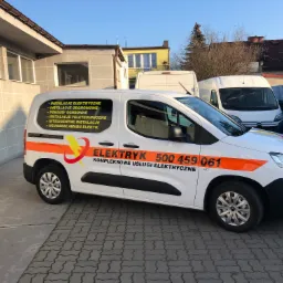 Biały samochód dostawczy z pomarańczowymi pasami i logiem elektryka, zaparkowany na betonowym podjeździe przed budynkiem. Na boku pojazdu widoczne wypisane usługi: instalacje elektryczne, odgromowe...