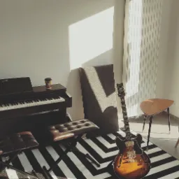 Jasna sala z pianinem Casio, gitarą elektryczną typu hollow body, pufą ze skóry, stolikiem i fotelem na tle czarno-białego dywanu w pasy; na pianinie stoi kubek z kawą.