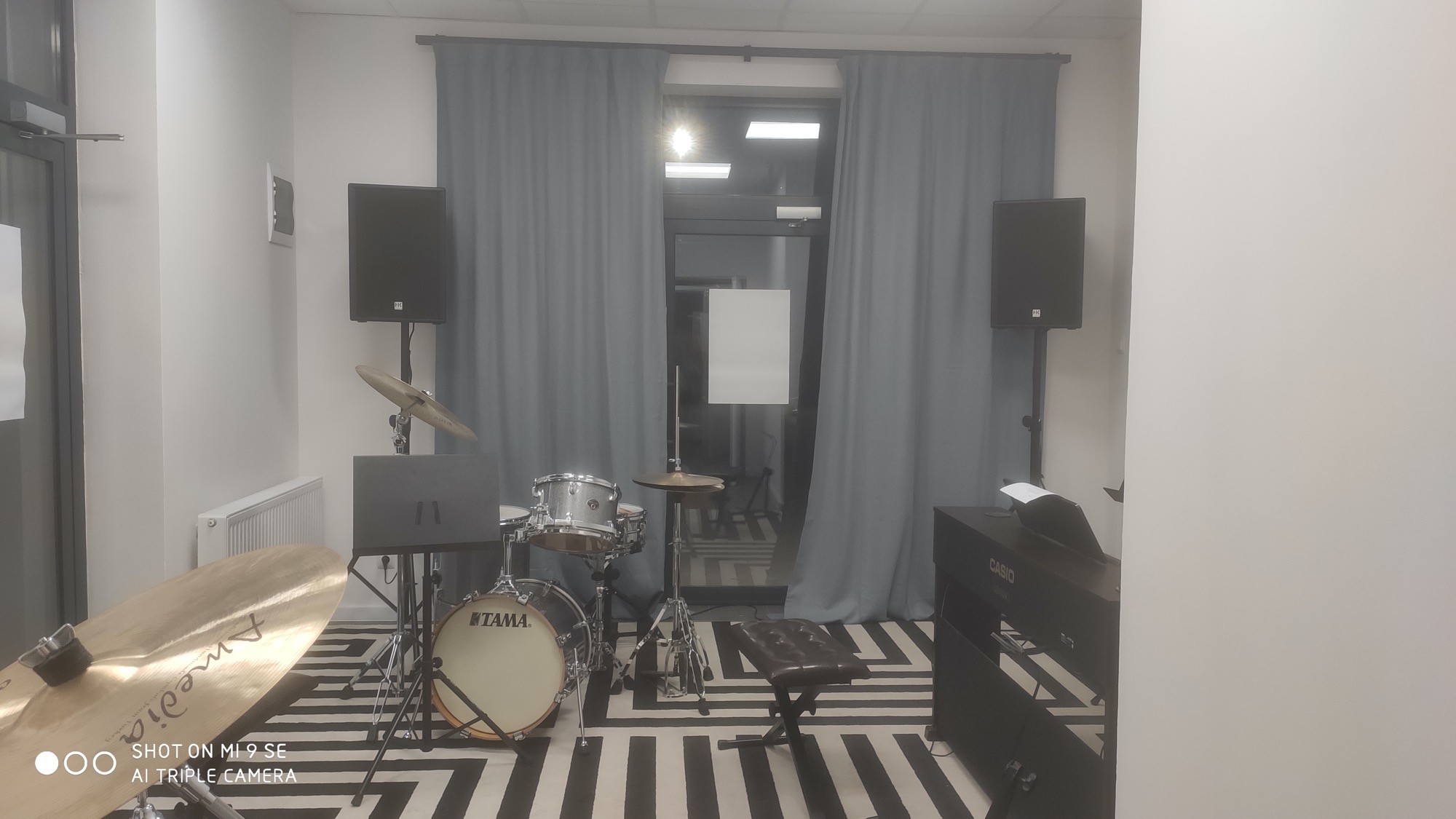 Sala z instrumentami muzycznymi: perkusja Tama, keyboard Casio, talerze Amedia, wzmacniacze HK Audio, laptop na statywie, krzesło i okno z zasłonami na tle dywanu w geometryczne wzory.