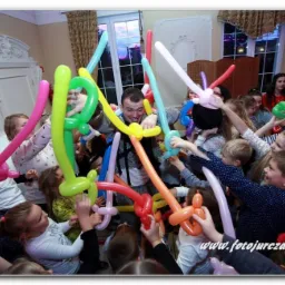 Animator w akcji: mężczyzna otoczony rozradowanymi dziećmi, prezentujący balony modelowane w kształty zwierząt i mieczy podczas imprezy urodzinowej.