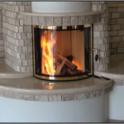 Otwarty palenisko z płomieniami, obudowane jasnym kamieniem i wykończone złotymi detalami, z kompletem akcesoriów kominkowych po lewej stronie i dekoracją roślinną na ścianie po prawej.