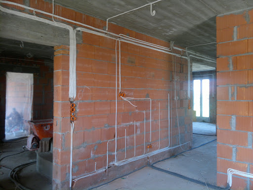 Instalacja elektryczna w surowym stanie: białe przewody prowadzone po ceglanych ścianach, widoczne puszki instalacyjne i betonowy sufit, w tle wózek budowlany.