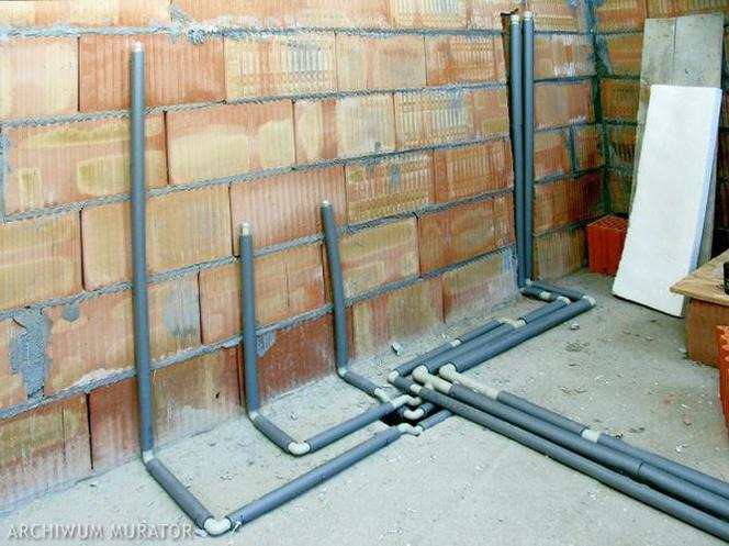 Szare rury instalacyjne wystające ze ściany z czerwonej cegły dziurawki, połączone kolankami na podłodze, widok z bliska na surową instalację.