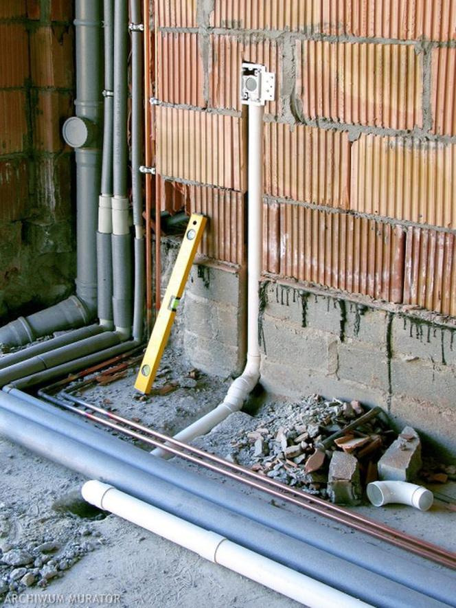 Instalacja elektryczna w trakcie montażu: widoczne puszki, rury PCV, poziomica i fragmenty cegieł na surowej ścianie.