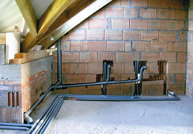 Surowe wnętrze z widocznymi elementami instalacji wodnej: szare rury prowadzone po ścianie z cegły dziurawki i wzdłuż podłogi, drewniana konstrukcja dachu w tle.