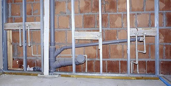 Szare rury kanalizacyjne w trakcie montażu w ścianie z cegieł dziurawki, widoczne metalowe profile konstrukcyjne i drewniane elementy mocujące.