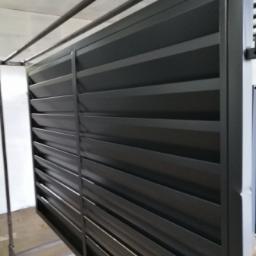 Prasmet Bieniek i Koniarczyk Sp.J - Nowoczesne, metalowe ogrodzenie lamelowe w kolorze grafitowym, zawieszone na metalowym stelażu w hali produkcyjnej przed montażem.