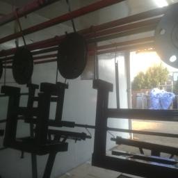 Prasmet Bieniek i Koniarczyk Sp.J - Czarne, metalowe elementy konstrukcyjne zawieszone na hakach w pomieszczeniu warsztatowym, prawdopodobnie po procesie malowania, z widocznym fragmentem podwórza i roślinności w tle.