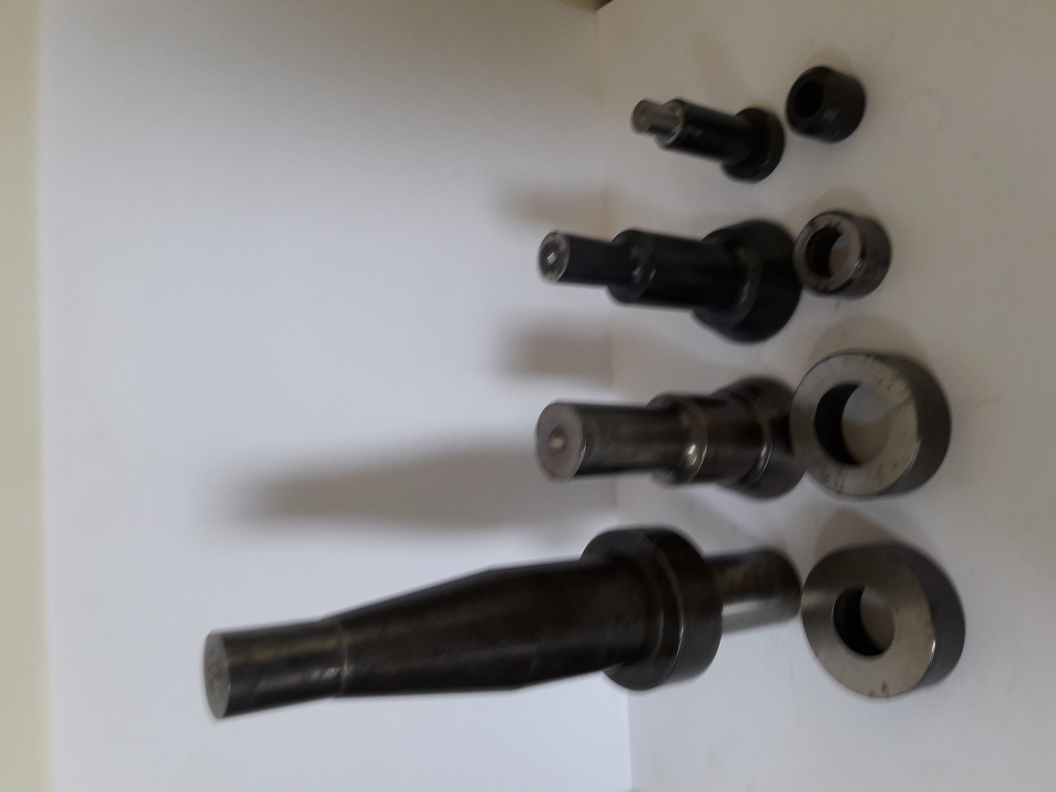 Różnej wielkości metalowe trzpienie i pierścienie ułożone w rzędzie na białym tle, widoczne ślady obróbki.