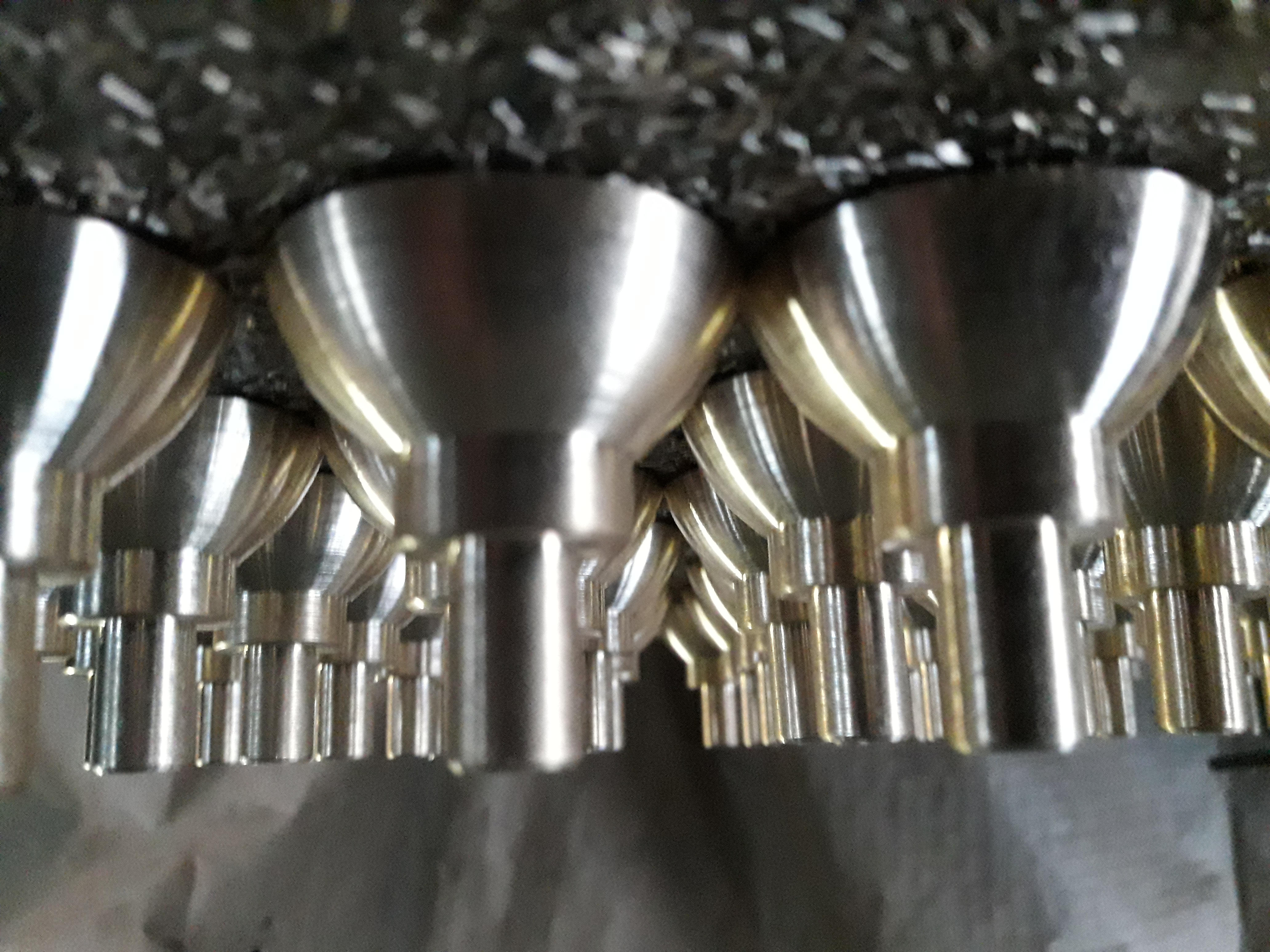 Rząd metalowych, stożkowych elementów z cylindrycznymi podstawami, prawdopodobnie części maszyn, ułożonych na tle metalowych opiłków.