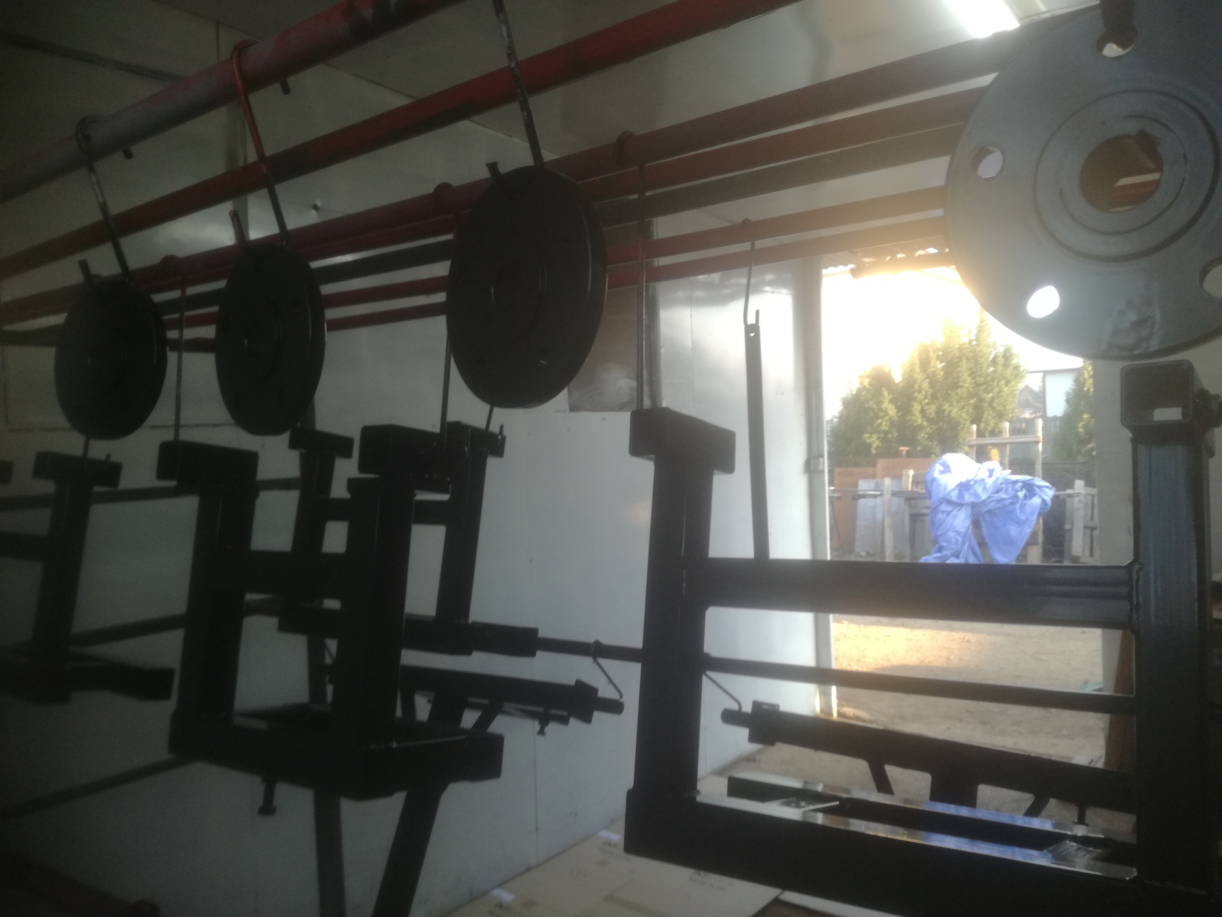 Czarne, metalowe elementy konstrukcyjne zawieszone na hakach w pomieszczeniu warsztatowym, prawdopodobnie po procesie malowania, z widocznym fragmentem podwórza i roślinności w tle.