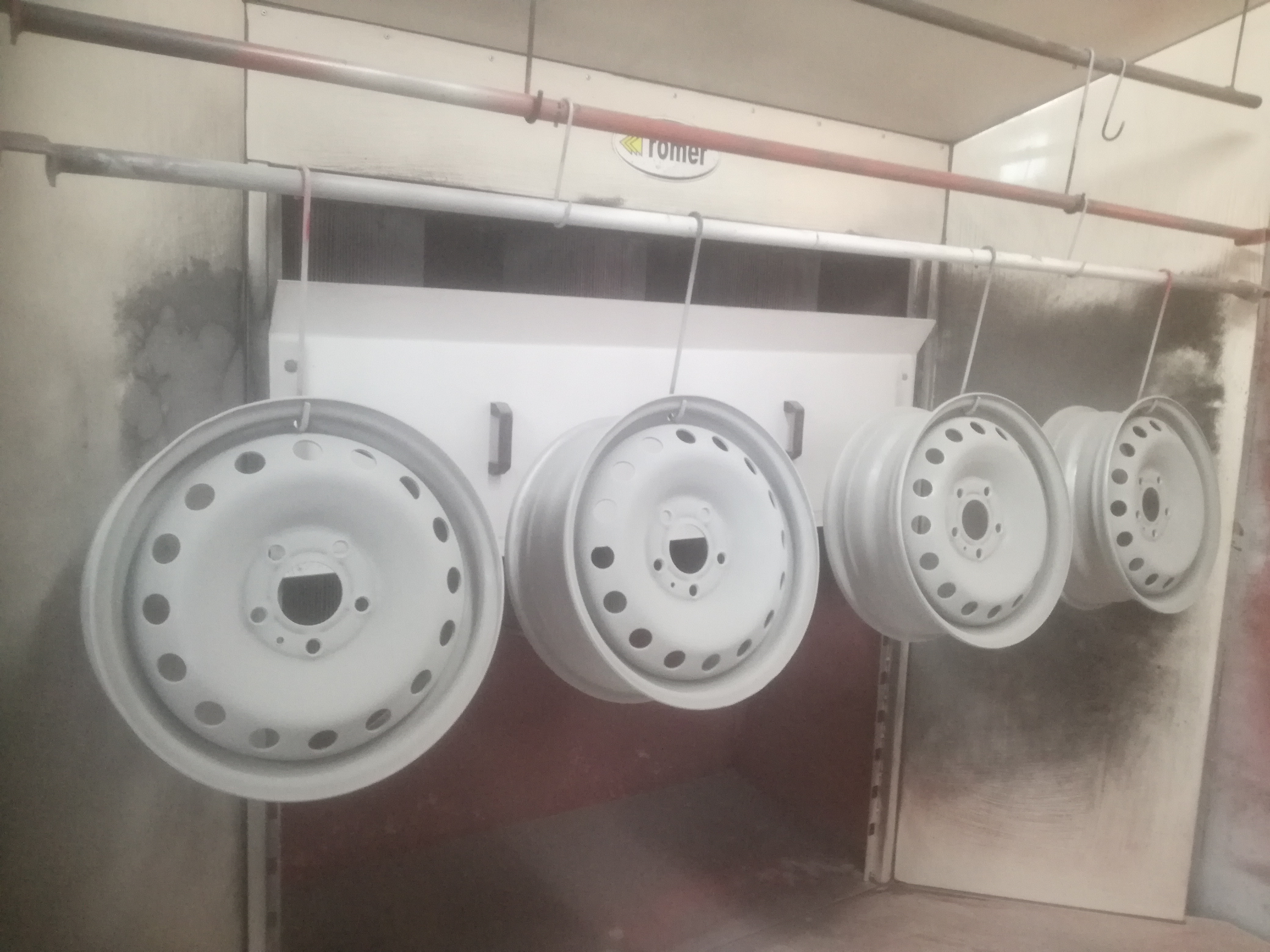 Cztery metalowe felgi samochodowe zawieszone na hakach w kabinie lakierniczej, po nałożeniu białej farby proszkowej.