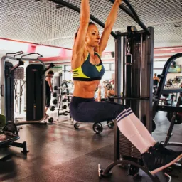 Kobieta w sportowym stroju wykonuje podciąganie na drążku w siłowni, widoczne napięte mięśnie brzucha i ramion, na tle inne urządzenia do ćwiczeń i osoby trenujące.