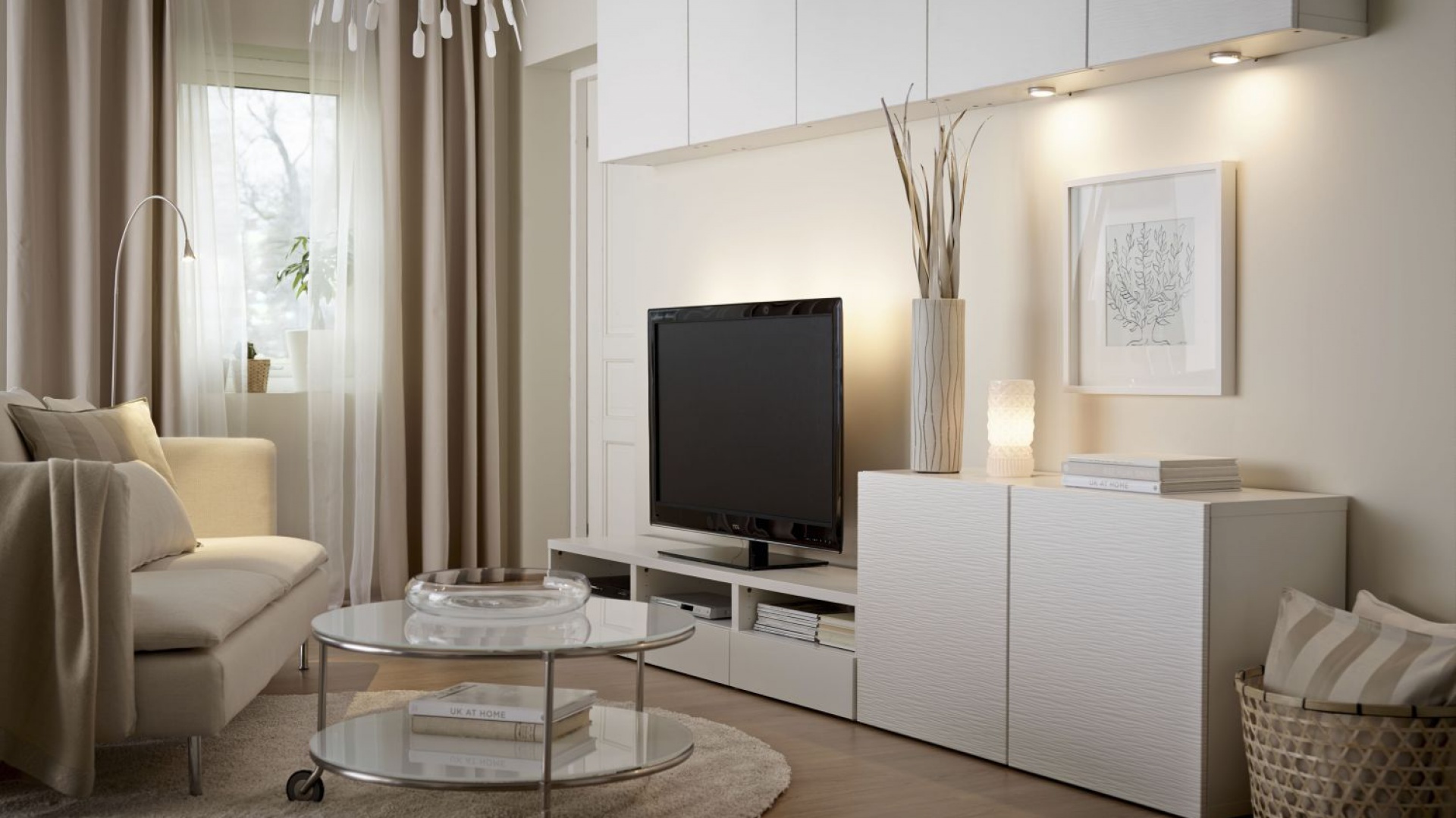 Jasny salon z beżową sofą, szklanym stolikiem kawowym i białą szafką RTV z telewizorem podwieszaną pod sufitem, dekoracje w postaci wazonu z suchymi gałązkami i obrazka w ramce.