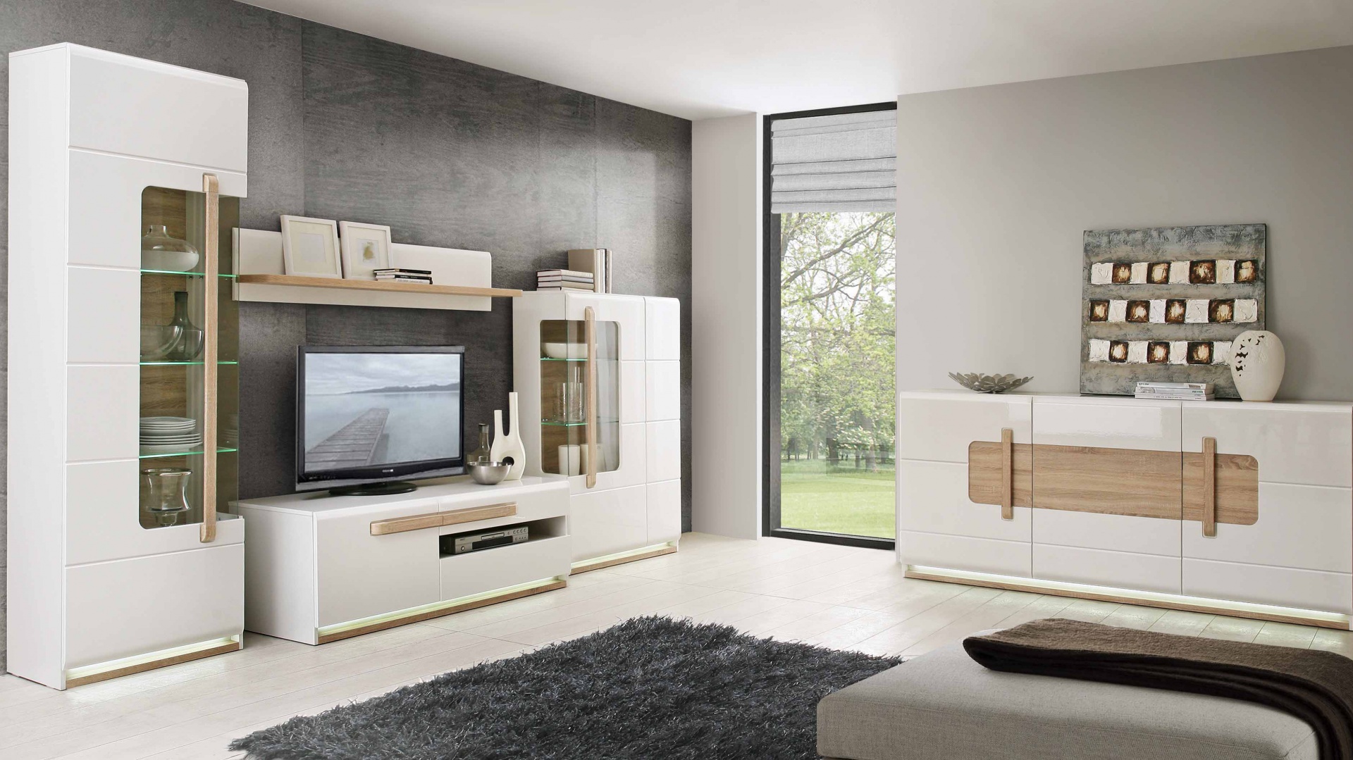Nowoczesny zestaw mebli do salonu w kolorze białym z drewnianymi akcentami i podświetleniem LED, na tle szarej ściany i okna z widokiem na zieleń