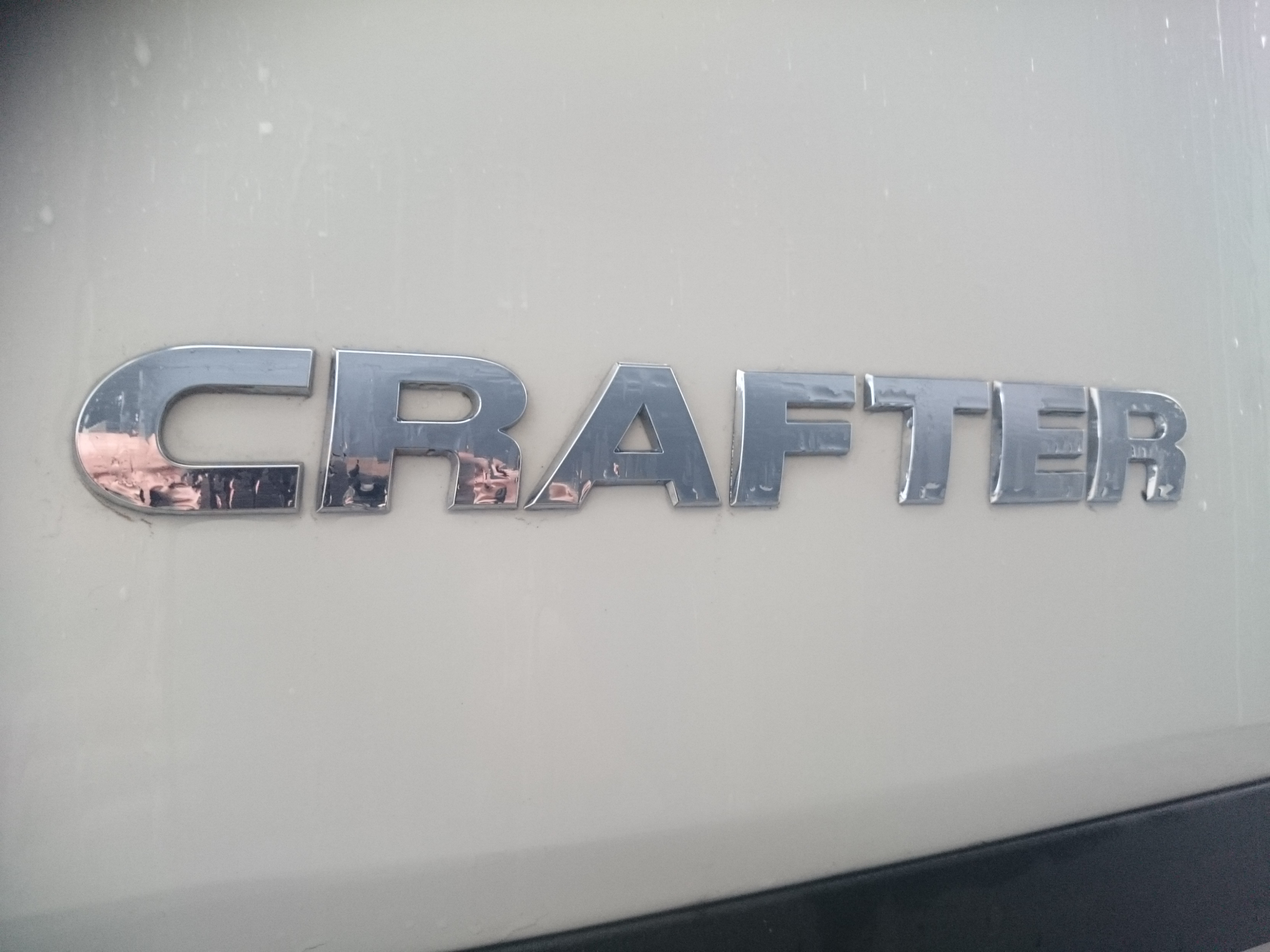 Chromowany napis 'CRAFTER' na boku jasnego, prawdopodobnie białego, brudnego samochodu dostawczego.