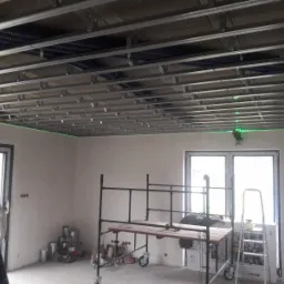 Montaż stelaża sufitowego z profili metalowych, widoczny laser krzyżowy wyznaczający poziom, rusztowanie robocze i drabina w pomieszczeniu w trakcie remontu.