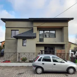 Dwukondygnacyjny dom z elewacją w odcieniach szarości, z czarną dach&oacute;wką i stolarką okienną, widziany z perspektywy ulicy, z zaparkowanym srebrnym samochodem osobowym i materiałami budowlanymi za...