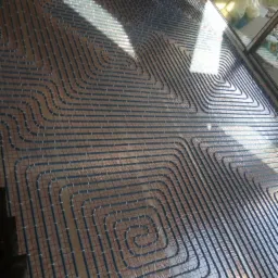 Rozłożone rury ogrzewania podłogowego na folii izolacyjnej z widoczną siatką montażową, oświetlone naturalnym światłem wpadającym przez okno.