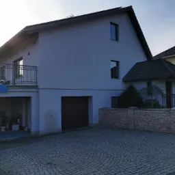 Jasnoszary dom jednorodzinny z balkonem i garażem, widok z brukowanego podjazdu w słoneczny dzień, elewacja po malowaniu.