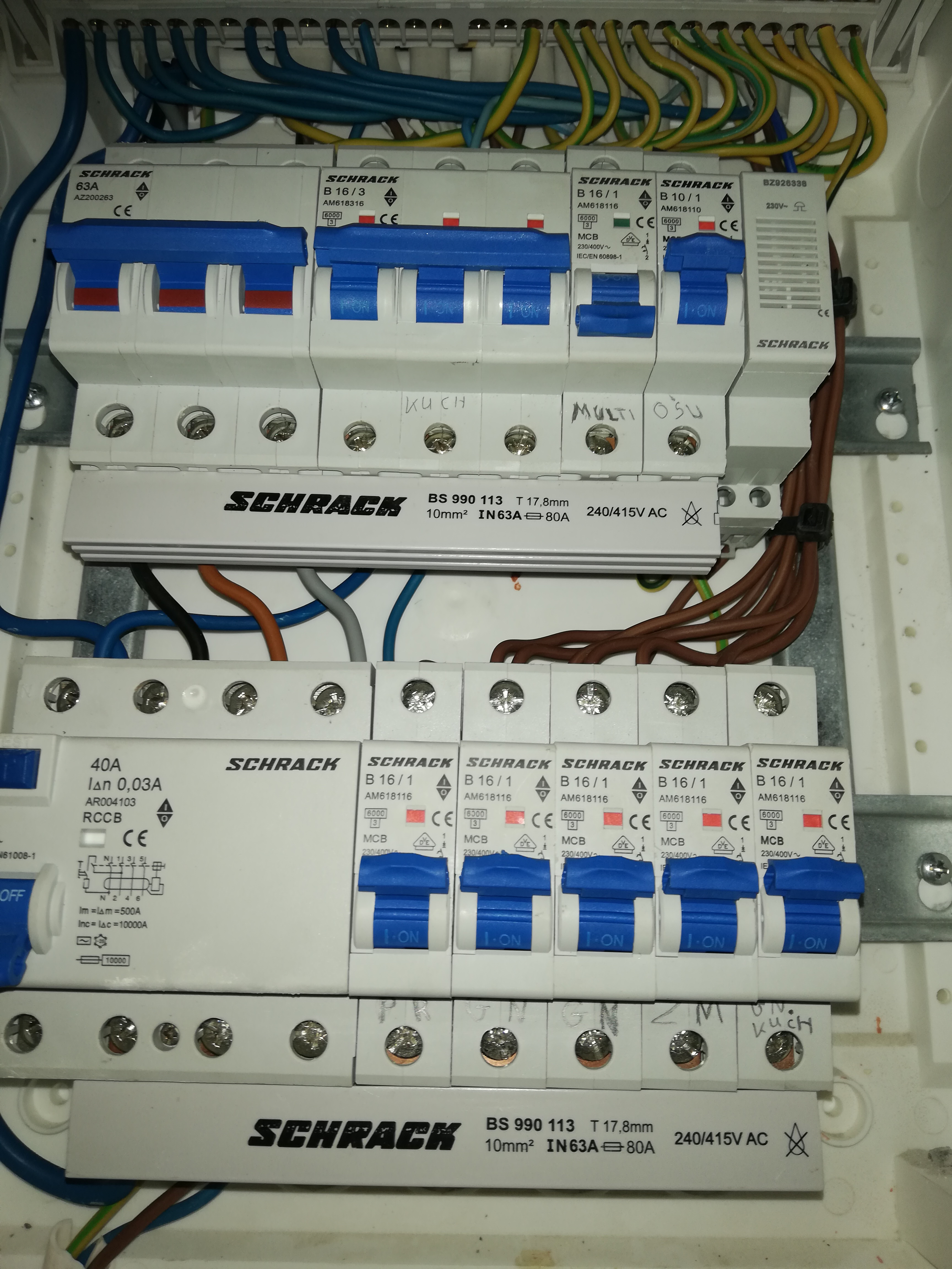 Rozdzielnica elektryczna z zainstalowanymi bezpiecznikami marki Schrack, widoczne przewody i oznaczenia obwodów, w tym 'Kuch' i 'Multi Osu'.