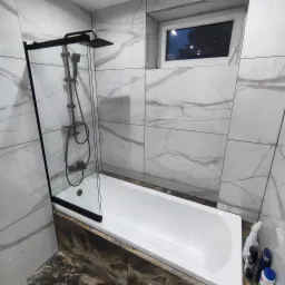 Nowoczesna łazienka z wanną obudowaną płytkami imitującymi drewno, przeszklonym parawanem nawannowym z czarną armaturą i ścianami wyłożonymi marmurowymi płytkami. Widoczne okno z widokiem na drzewa.