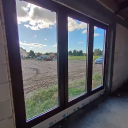 Trzyczęściowe okno z brązowymi ramami, widok na plac budowy, dom i samochód w tle, folia ochronna na dole ramy.