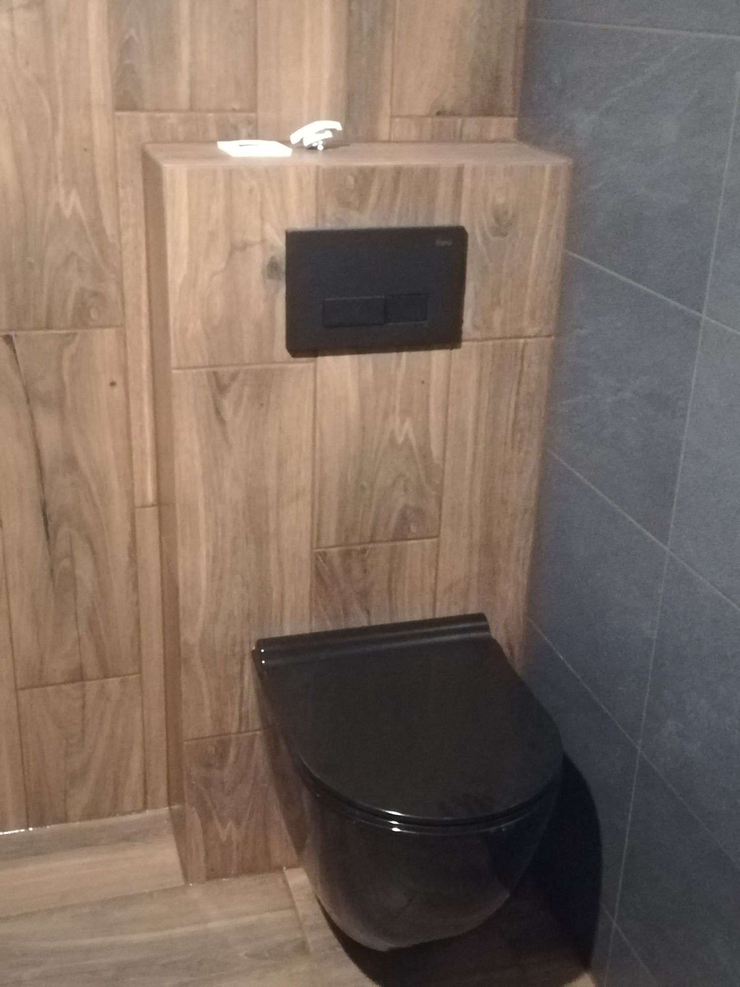 Czarna, wisząca toaleta ze spłuczką w zabudowie z płytek imitujących drewno, obok ściana wyłożona szarymi płytkami.