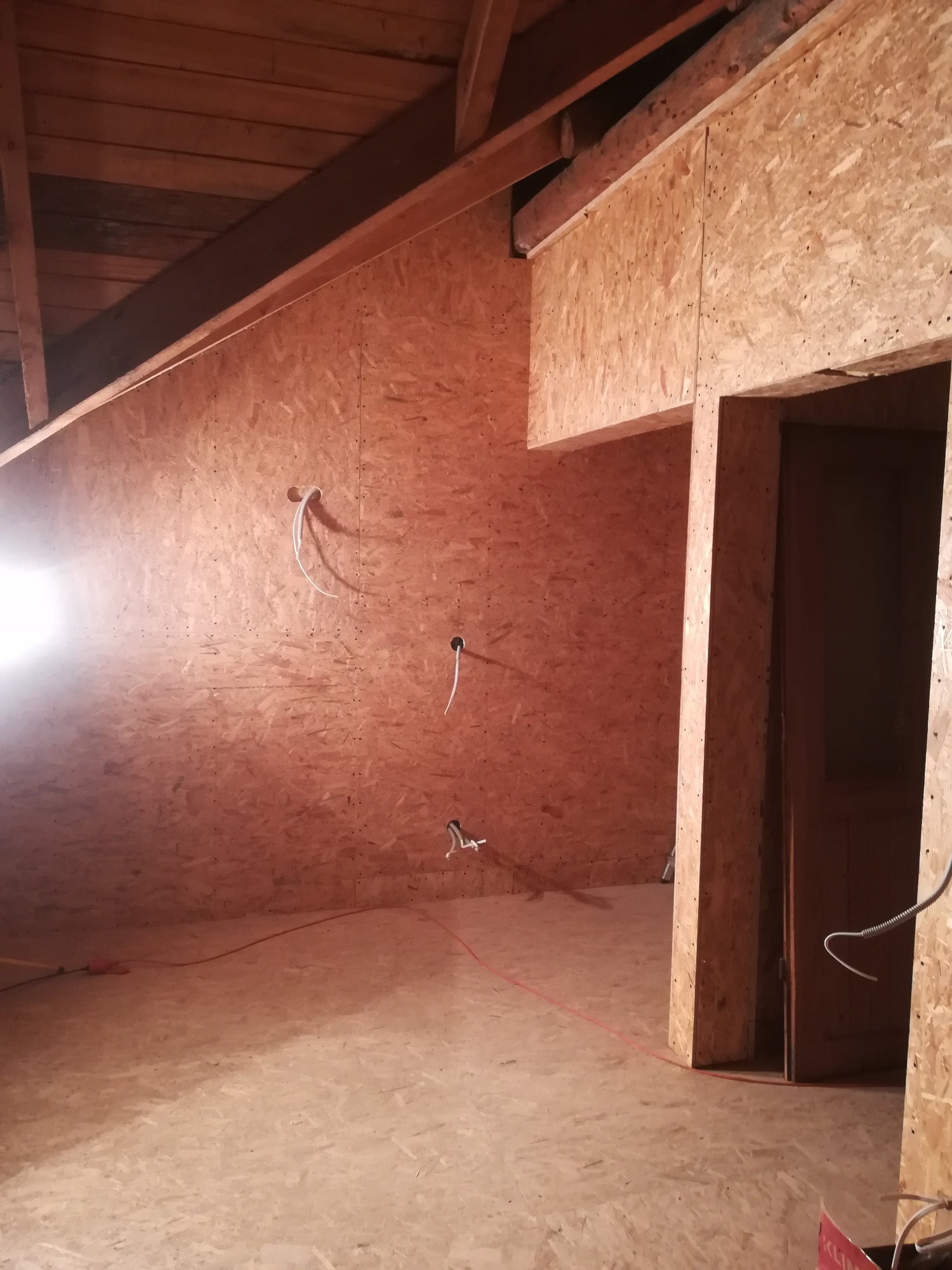 Poddasze wyłożone płytami OSB, widoczne kable elektryczne wystające ze ściany, drewniana konstrukcja dachu i rama drzwiowa, oświetlenie robocze po lewej stronie.