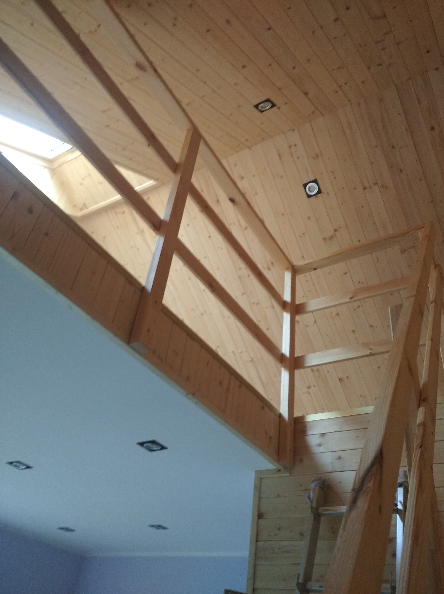 Drewniane wykończenie poddasza z drewnianą balustradą i schodami, widoczne oświetlenie punktowe w suficie i skos dachu z oknem.