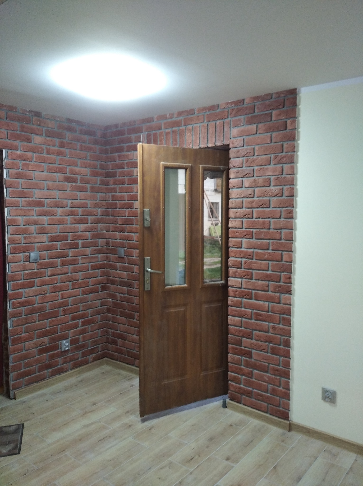 Wnętrze z drewnianymi panelami podłogowymi, ścianą wyłożoną dekoracyjną cegłą i drewnianymi drzwiami z przeszkleniami.