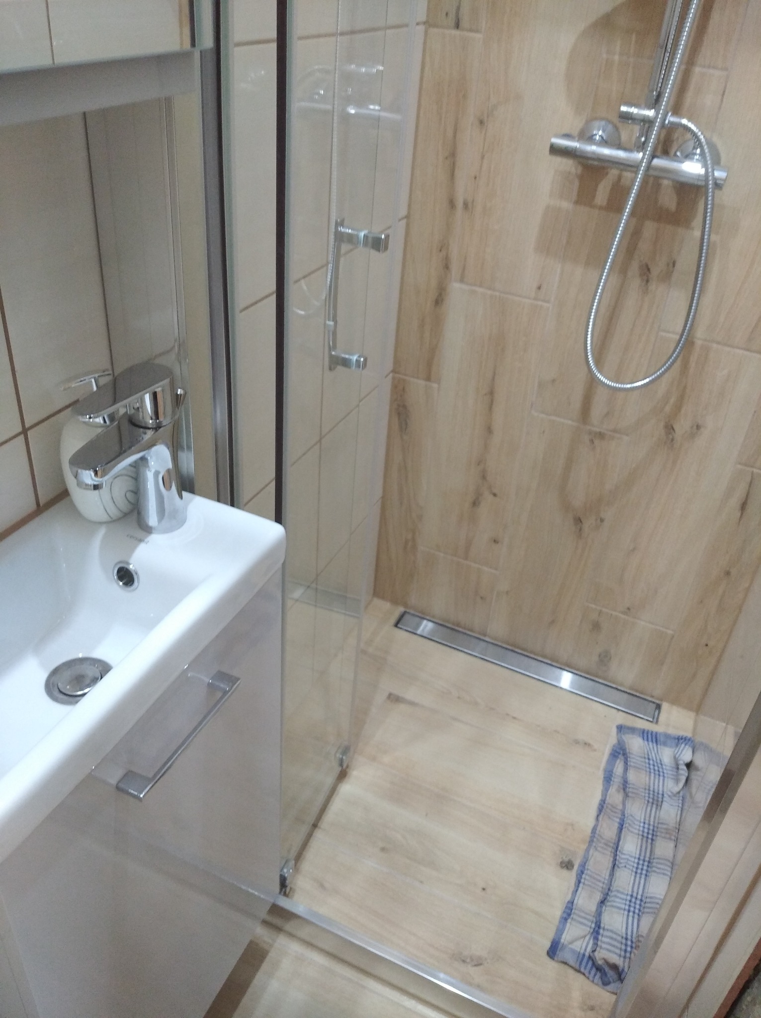 Nowoczesna łazienka z kabiną prysznicową typu walk-in, drewnopodobnymi płytkami na ścianach i podłodze, minimalistyczną umywalką z szafką oraz odpływem liniowym; na podłodze leży niebieska szmatka.