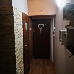 AW - Wnętrze przedpokoju z dekoracyjną ścianą z kamienia, drewnianymi drzwiami wejściowymi z ozdobnym wieńcem w kształcie serca, szafkami nad drzwiami i oświetleniem sufitowym.