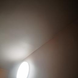 AW - Okrągła lampa sufitowa LED, zamontowana na białym suficie, oświetlająca fragment ściany w odcieniach beżu, widok z dołu.