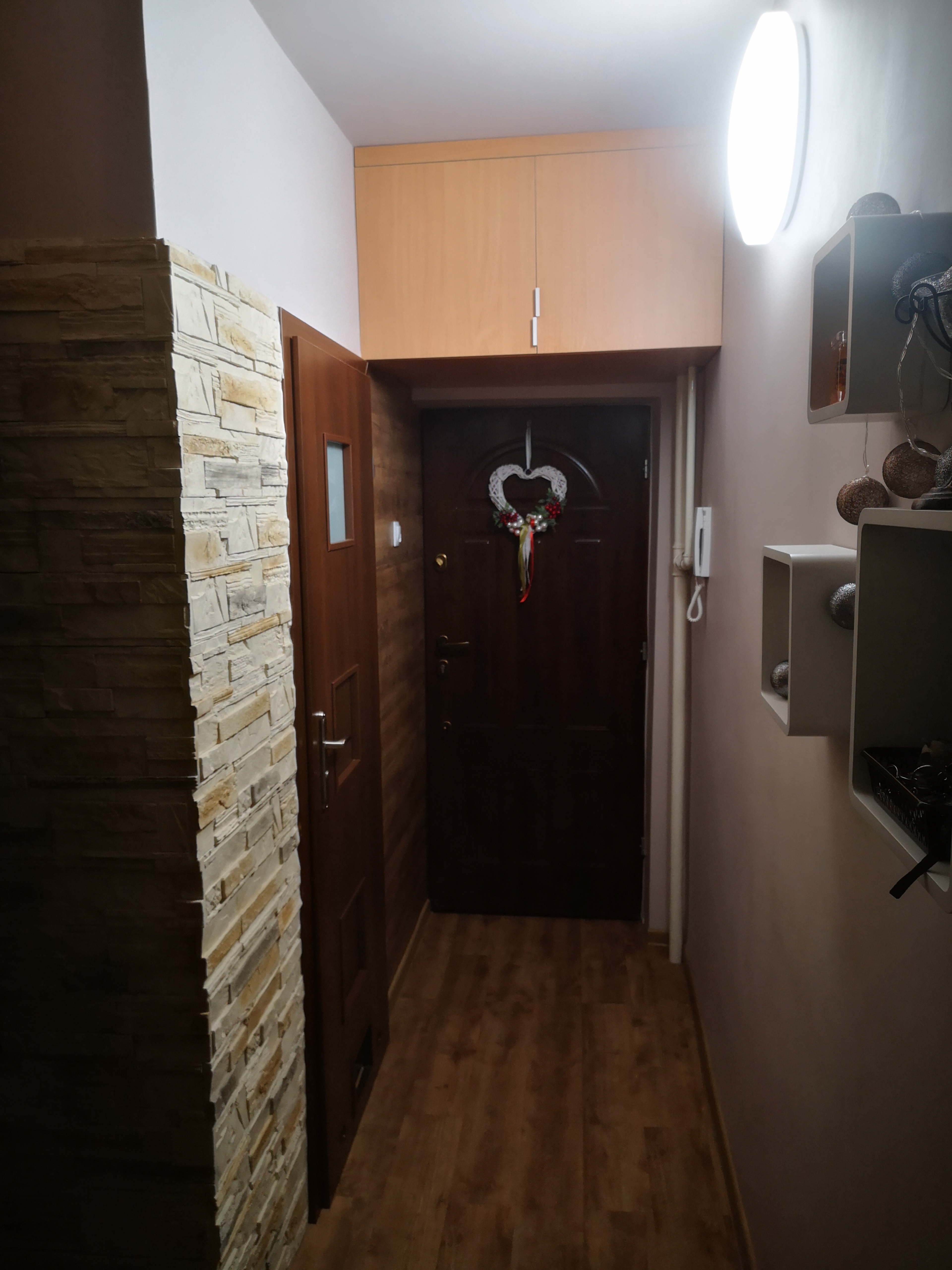Wnętrze przedpokoju z dekoracyjną ścianą z kamienia, drewnianymi drzwiami wejściowymi z ozdobnym wieńcem w kształcie serca, szafkami nad drzwiami i oświetleniem sufitowym.
