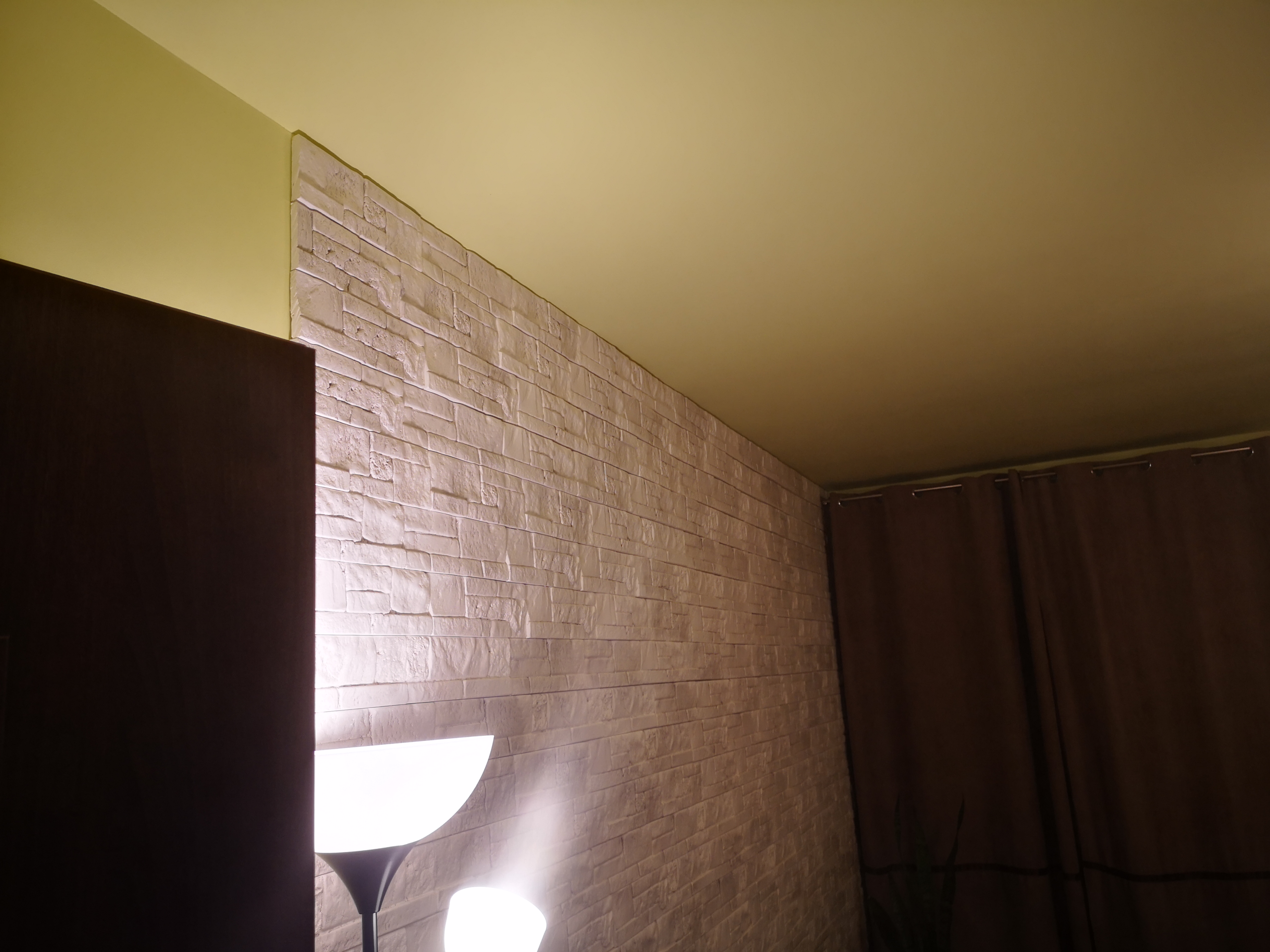Ściana wykończona dekoracyjną płytą karton gips imitującą cegłę, oświetlona przez lampę podłogową, widok z dołu.