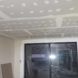 Sufit z płyt kartonowo-gipsowych w trakcie montażu, widoczne łączenia szpachlowane, przewody elektryczne wystające z otworów, ściany przygotowane do dalszych prac wykończeniowych, okno z roletami...