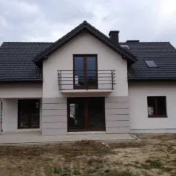 Nowoczesny dom jednorodzinny z białymi ścianami i ciemną dachówką, balkon z czarną balustradą, elewacja częściowo wykończona szarymi panelami, widoczne okna z brązowymi ramami, teren wokół domu...