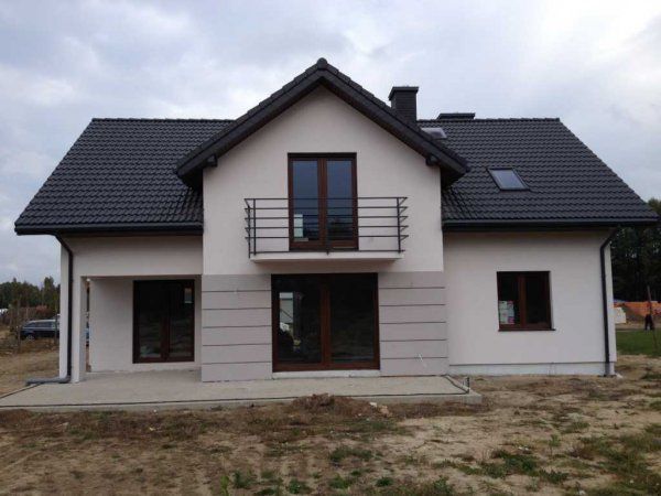 Nowoczesny dom jednorodzinny z białymi ścianami i ciemną dachówką, balkon z czarną balustradą, elewacja częściowo wykończona szarymi panelami, widoczne okna z brązowymi ramami, teren wokół domu...