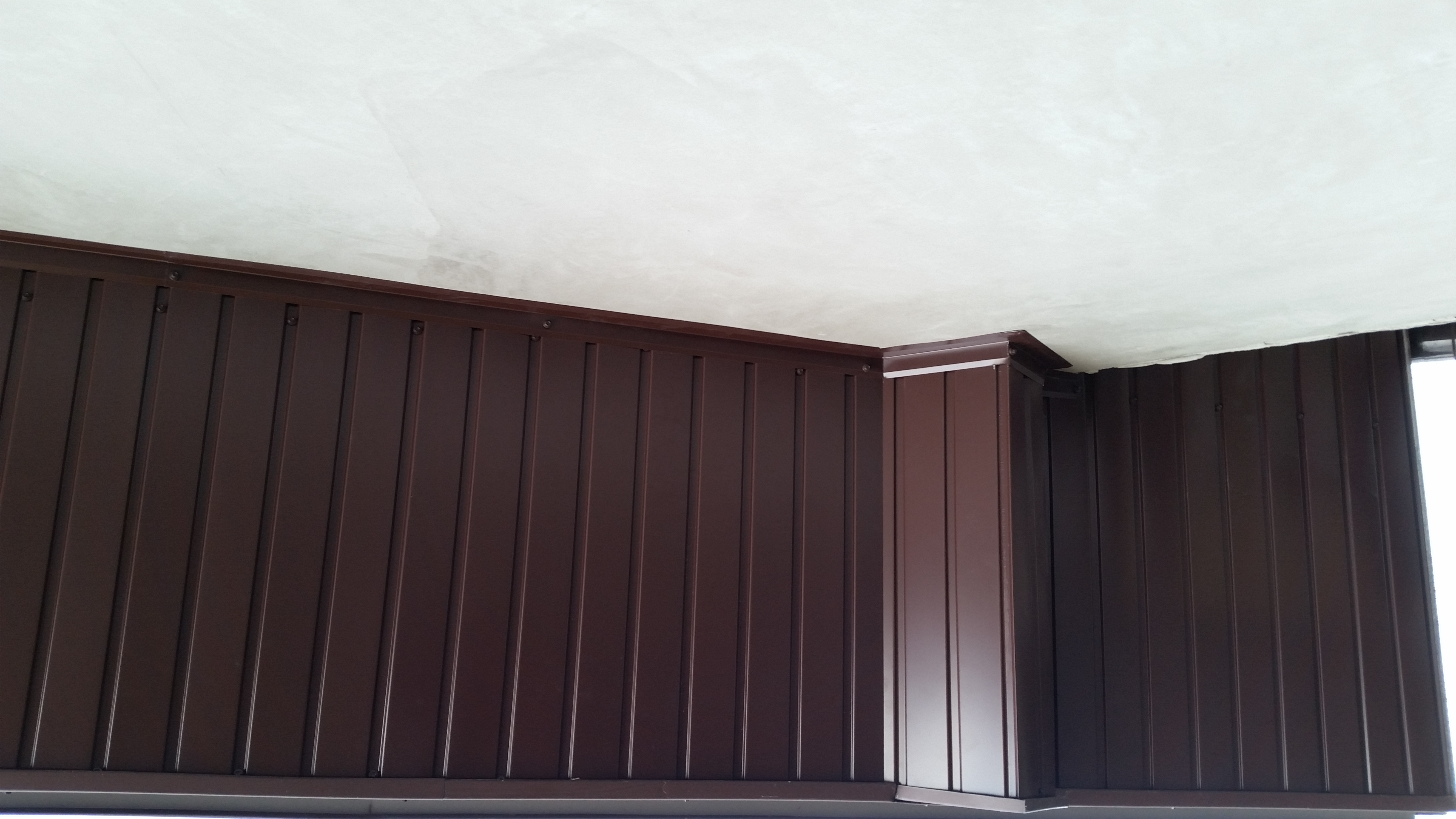 Brązowa blacha trapezowa na zadaszeniu, widok od dołu, mocowana na śruby, z widocznym fragmentem betonowego sufitu i okna.