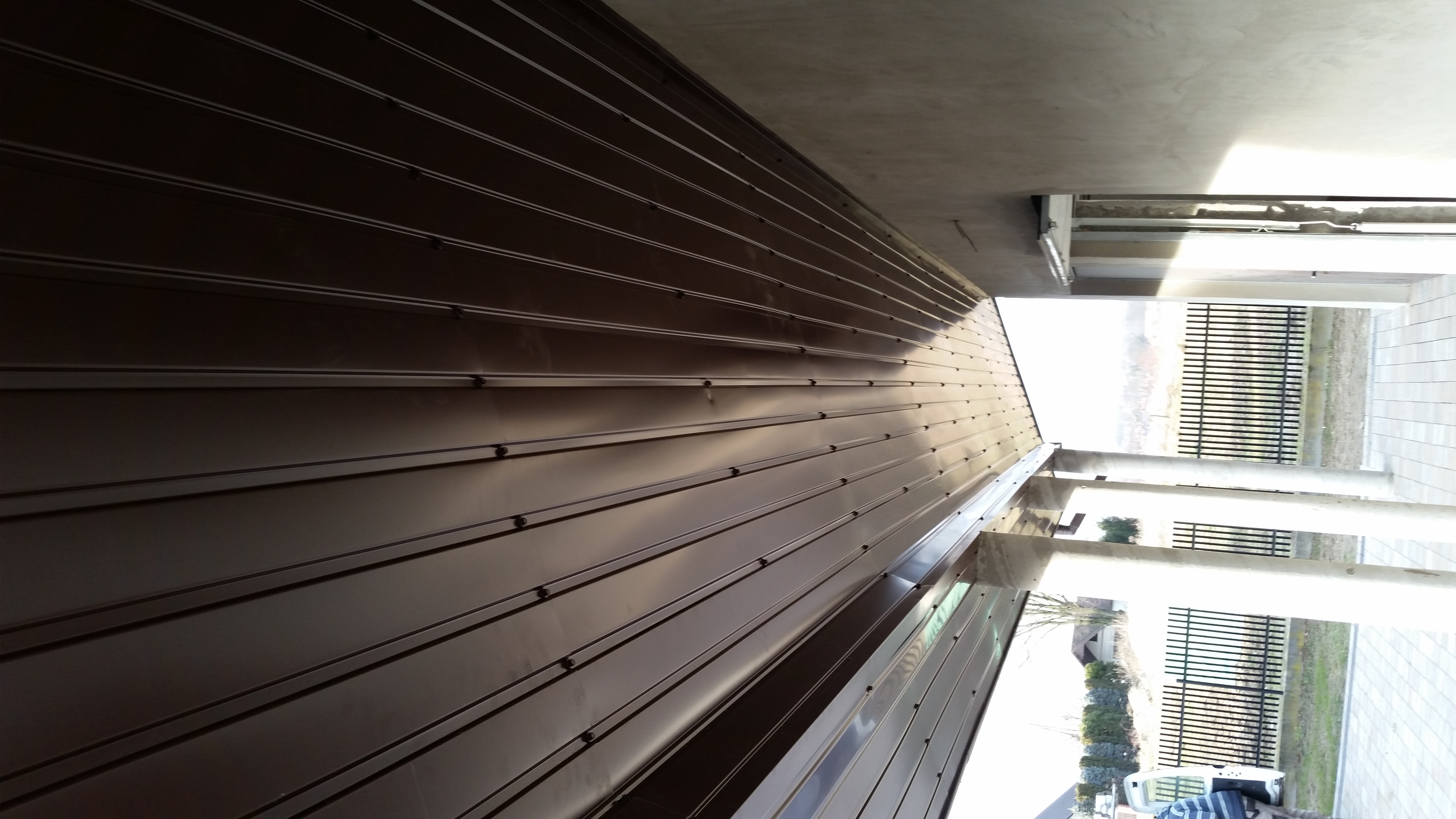 Nowe, brązowe pokrycie dachu z blachy trapezowej, z widocznymi śrubami mocującymi, sfotografowane pod kątem, z fragmentem betonowej konstrukcji budynku w tle.