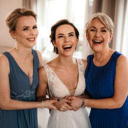 ACfotografia fotografia ślubna Gdańsk Trójmiasto - Panna młoda w białej sukni ślubnej, trzymająca dłonie z dwiema uśmiechniętymi kobietami w niebieskich sukienkach, śmieją się radośnie w jasnym wnętrzu.