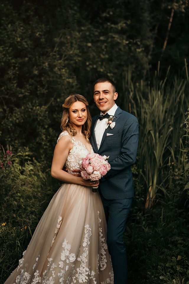 Uśmiechnięta para młoda w plenerze: panna młoda w sukni z koronkowymi aplikacjami trzyma bukiet różowych piwonii, pan młody w granatowym garniturze z muchą, na tle zieleni.