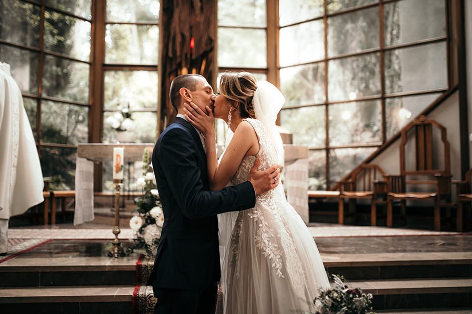 Nowożeńcy w kościele, pocałunek po ceremonii, ona w białej sukni z koronkowymi aplikacjami, on w granatowym garniturze, w tle drewniany ołtarz i okna z widokiem na zieleń.