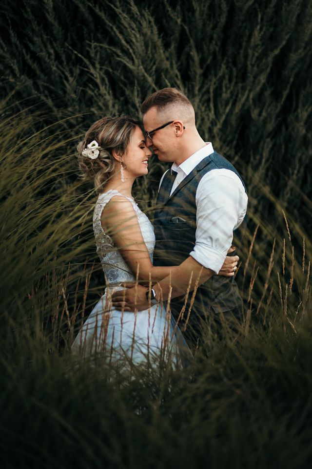 Para młoda obejmująca się w wysokiej trawie, panna młoda z kwiatami we włosach i delikatnymi kolczykami, pan młody w okularach i kamizelce.