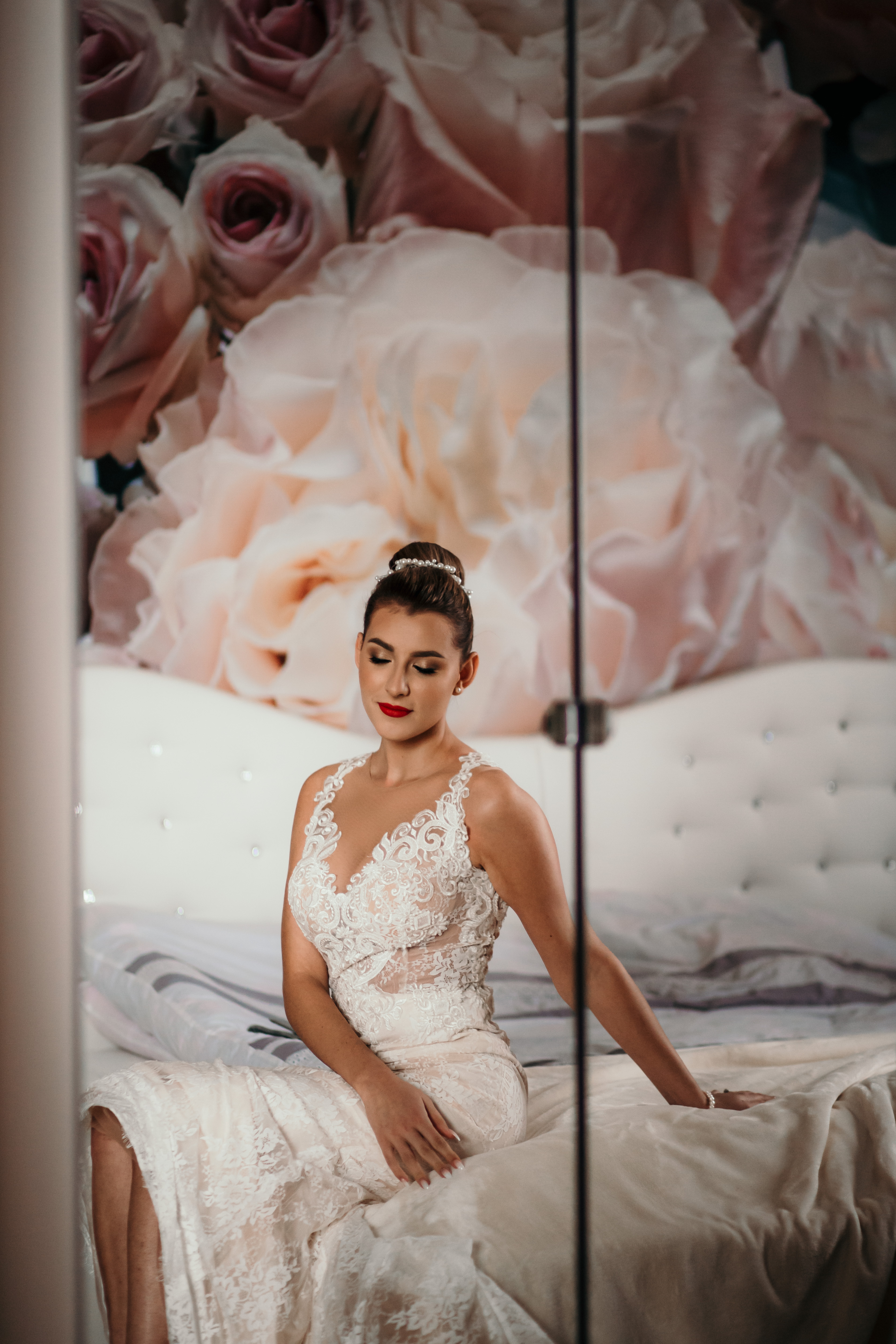 Panna młoda w koronkowej sukni siedzi na łóżku z kwiatowym tłem, z zamkniętymi oczami i czerwonymi ustami.