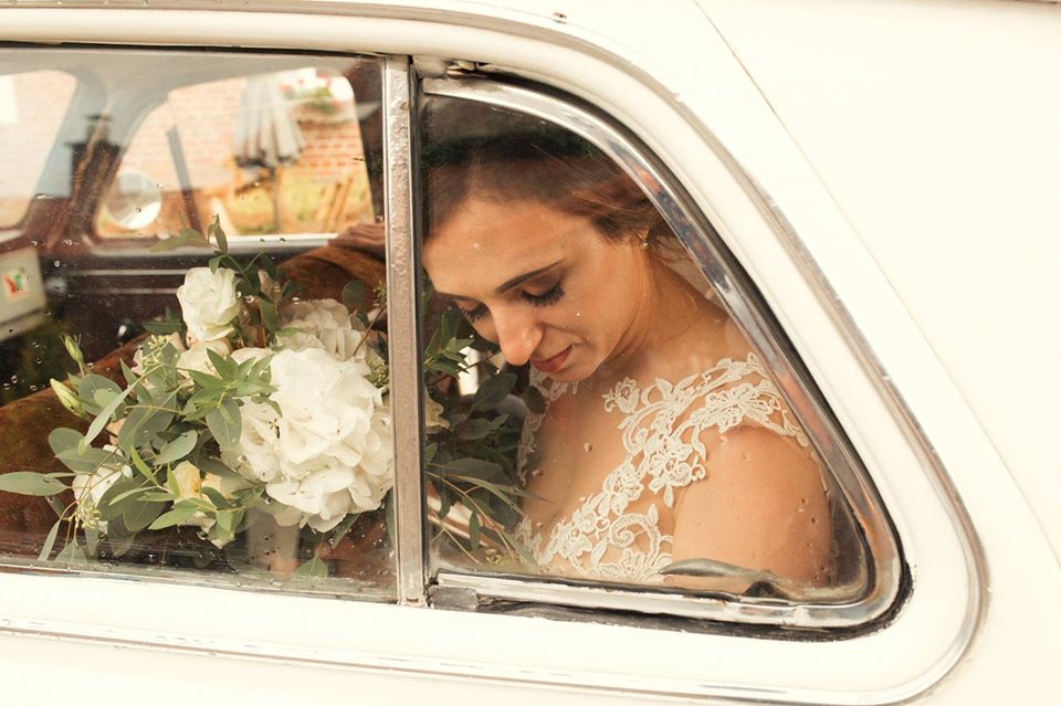 Panna młoda w koronkowej sukni spogląda w dół, siedząc w samochodzie retro z bukietem białych kwiatów, krople deszczu na szybie.