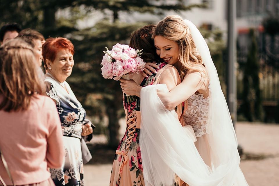Panna młoda w koronkowej sukni ślubnej i welonie obejmuje kobietę z bukietem różowych piwonii, w tle uśmiechnięte osoby.